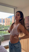 A woman wearing a light pink lace bralette and blue jeans stands in a modern room with a large window overlooking a cityscape.